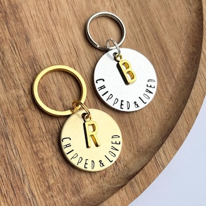 Könnte beinhalten: Zwei runde Schlüsselanhänger mit den Worten "CHIPPED & LOVED" in schwarzer Schrift. Ein Schlüsselanhänger ist goldfarben mit einem goldenen "R"-Anhänger, der andere ist silberfarben mit einem goldenen "B"-Anhänger. Beide Schlüsselanhänger haben einen silbernen oder goldenen Ring.