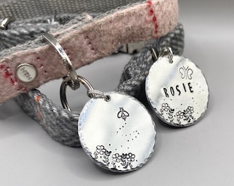 May include: Two silver dog tags with engraved designs. One tag has the name "Rosie" with a butterfly and floral designs. The other tag has a bee and floral designs.