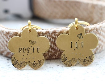 May include: Two gold flower-shaped dog tags with engraved names, Rosie and Rae, and floral designs. The tags have a small bee and butterfly charm.