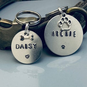 Könnte beinhalten: Zwei silberne Hundemarken mit den eingravierten Namen "Daisy" und "Archie". Die Marken haben einen Knochen- und einen Pfotenabdruck-Anhänger. Die Marken sind an einem braunen Lederhundekragen befestigt.