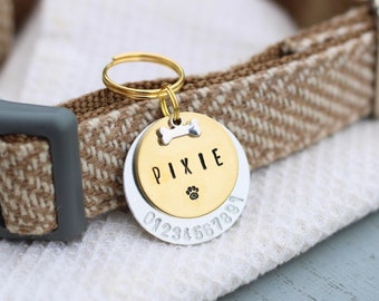 May include: Gold and silver dog tag with the name "Pixie" engraved on it. The tag also has a bone and paw print design. The tag is attached to a brown and white dog collar.