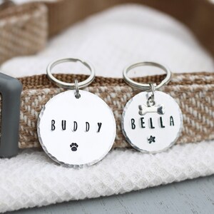 K&ouml;nnte beinhalten: Zwei silberne Hundemarken mit den personalisierten Namen "Buddy" und "Bella". Die Marken sind an einem braunen und wei&szlig;en Hundehalsband befestigt.