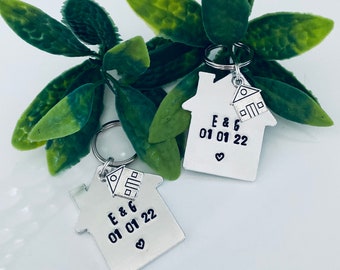 May include: Two silver house-shaped keychains with a smaller house charm attached. The keychains are engraved with "E & G 01 01 22" and a heart symbol.