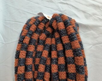 May include: A pair of handmade mittens in a checkerboard pattern of orange and dark gray. The mittens are made of a thick, textured yarn, and are likely designed for warmth. A small tag is attached to one of the mittens.