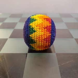 May include: A rainbow crocheted ball with a blue, green, yellow, orange, and red pattern. The ball is sitting on a black and white chequered surface.