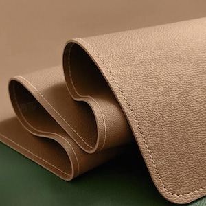 Puede incluir: Un tapete de escritorio de cuero enrollado, de color marrón claro, con costuras blancas en los bordes. El tapete descansa parcialmente sobre una superficie verde oscuro, creando un contraste de color y textura. El cuero tiene una sutil textura granulada.