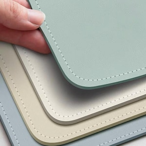 May include: Three rectangular placemats in shades of blue and beige are stacked. Each placemat has rounded corners and a stitched border. A hand holds the top placemat, which is light blue.