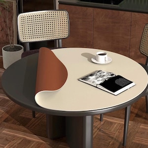 May include: A round table with a two-tone surface, featuring a light beige top and a dark gray base. A brown mat is partially rolled back, revealing the gray surface. A cup of coffee and an open book rest on the table. Two chairs with woven backs are in the background.