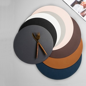 Peut inclure: Ensemble de sept sets de table ronds en cuir de différentes couleurs, dont gris, marron, beige et bleu. Les sets de table sont disposés en cercle avec une fourchette et un couteau dorés sur le dessus du set de table gris.