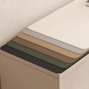 Puede incluir: Una pila de siete alfombrillas de escritorio de diferentes colores, en tonos beige, marrón, verde, negro, gris y blanco, sobre un armario beige claro con una puerta curva.