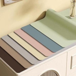 May include: A set of seven table mats in various shades of brown, beige, grey, pink, blue, and green. The mats are arranged in a stack, with the green mat on top. The mats are made of a smooth, textured material.