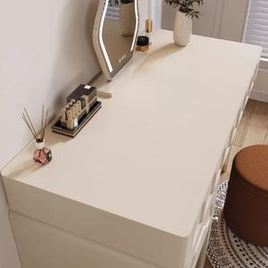 May include: A cream-colored vanity with a unique, illuminated mirror. The mirror has a wavy, organic shape with a bright white LED border. The vanity is topped with beauty products and a vase of flowers. A small brown stool sits nearby.