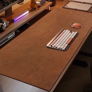 以下が含まれることがあります： 茶色のデスクで、大きな革のような表面があります。白と赤のキーが付いたレトロスタイルのキーボードがデスクにあり、ピンクのマウスもあります。モニターとデスクオーガナイザーが背景に表示され、装飾品があります。