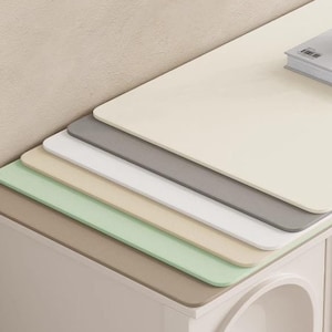 Może przedstawiać: Pięć różnych kolorowych podkładek biurkowych ułożonych jedna na drugiej. Podkładki są beżowe, jasnozielone, brązowe, białe i jasnoszare. Podkładki leżą na białym stole z wazą i książką na górze.