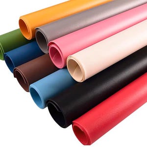 Op de afbeelding: Een verzameling opgerolde vellen gekleurd materiaal, waaronder tinten groen, blauw, bruin, roze, zwart en rood. De vellen zijn diagonaal gerangschikt, waarbij hun verschillende tinten en texturen worden getoond. Het materiaal lijkt flexibel en glad.