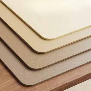 May include: A close-up of several rectangular placemats in varying shades of beige and tan. The placemats have rounded corners and a textured surface, with a wooden surface in the foreground.