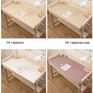 May include: Four different colored desk mats on a wooden desk. The mats are in the colors apricot, grayish tan, white, and lavender. Each mat is on a desk with a lamp and a book on it.