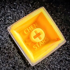 Pu&ograve; includere: Un keycap giallo con una cornice in plastica trasparente. Il keycap ha il testo "CHEN STARS" e un segno pi&ugrave; al centro.
