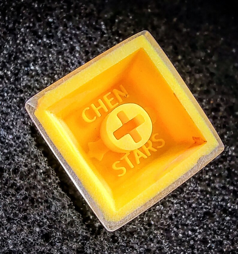 Pu&ograve; includere: Un keycap giallo con una cornice in plastica trasparente. Il keycap ha il testo "CHEN STARS" e un segno pi&ugrave; al centro.