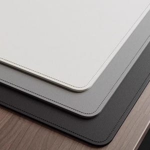 May include: Three leather desk mats in different shades of gray, from white to dark gray. The mats are stitched around the edges and are layered on top of each other.