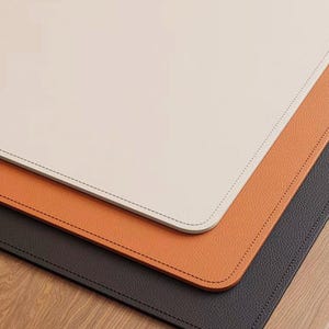 May include: A stack of three rectangular desk mats in cream, orange, and dark gray. Each mat has rounded corners and a stitched border. The mats are arranged on a wooden surface.