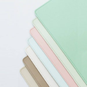 May include: A stack of seven colorful desk mats in various pastel shades, including light green, pink, blue, white, beige, and brown. The mats are arranged in a fan shape, with the light green mat at the top and the brown mat at the bottom.