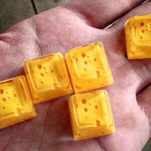 Pu&ograve; includere: Cinque keycap gialle a forma di cubetti di formaggio svizzero. Le keycap hanno un aspetto traslucido e sono disposte in fila su una mano.