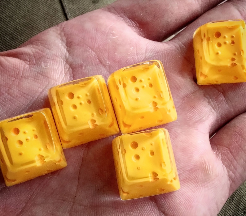 Pu&ograve; includere: Cinque keycap gialle a forma di cubetti di formaggio svizzero. Le keycap hanno un aspetto traslucido e sono disposte in fila su una mano.