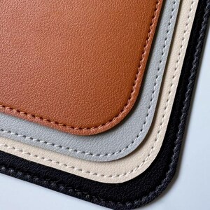 Könnte beinhalten: Nahaufnahme von vier Lederuntersetzern in verschiedenen Farben: Braun, Grau, Beige und Schwarz. Die Untersetzer haben einen genähten Rand und sind übereinander gestapelt.