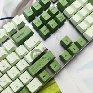 May include: A green and white mechanical keyboard with a mountain scene keycap. The keyboard has a silver frame and green and white keys with Japanese characters and English letters.