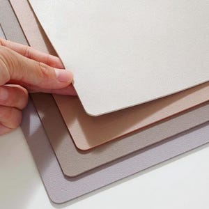 Puede incluir: Un juego de cinco manteles individuales en varios tonos de beige, gris y rosa. Los manteles individuales están hechos de un material texturizado que se asemeja al cuero.