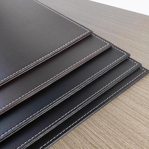 Pode incluir: Quatro tapetes de mesa de couro preto com costuras brancas ao redor das bordas. Os tapetes de mesa estão empilhados uns sobre os outros.