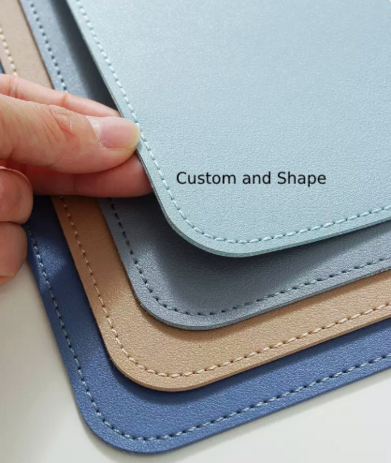 May include: Three leather desk mats in different shades of blue, beige, and light blue. The mats have stitched edges and are stacked on top of each other. The top mat has the text "Custom and Shape" printed on it.