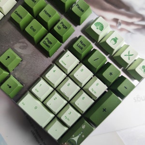 May include: A green and white mechanical keyboard with keycaps featuring various designs, including a fan, a girl with a flower, a sword, and a clover. The keyboard has a silver frame and green and white keys.