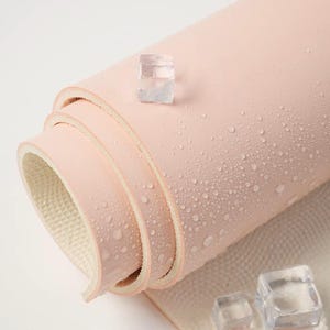 May include: A pink yoga mat rolled up with water droplets on the surface. The mat is made of a textured material and has a white underside. There are ice cubes on the white surface below the mat.