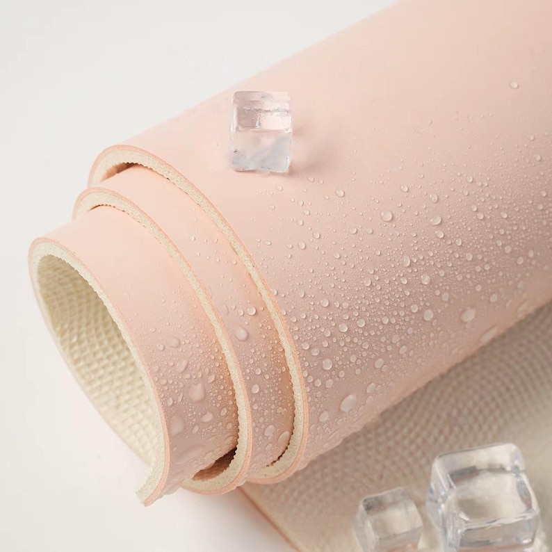 May include: A pink yoga mat rolled up with water droplets on the surface. The mat is made of a textured material and has a white underside. There are ice cubes on the white surface below the mat.