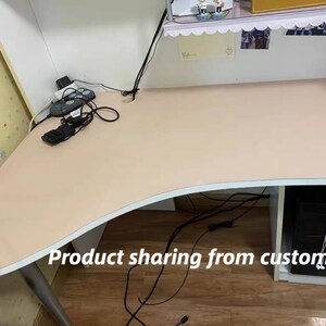 May include: A light pink desk with a curved edge and a white base. The desk has a power strip and cords on the surface. The desk is sitting on a wooden floor.