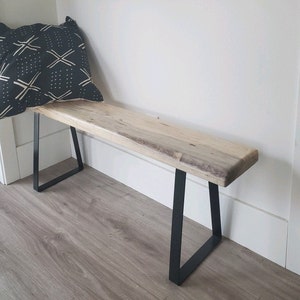 May include: A wooden bench with a live edge top and black metal A-frame legs. The bench is sitting on a wood floor.