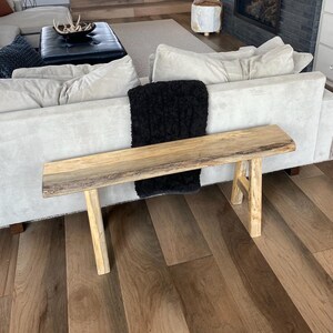 May include: A natural wood bench with a live edge top and two legs. The bench is in front of a white couch with a black throw blanket.