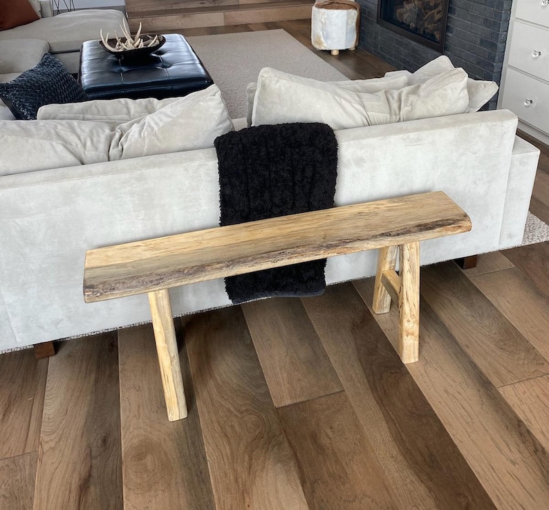 May include: A natural wood bench with a live edge top and two legs. The bench is in front of a white couch with a black throw blanket.
