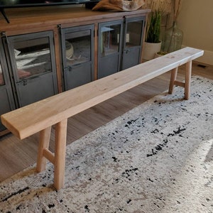 May include: A light wood bench with a simple design and two legs on each end. The bench is sitting on a patterned rug in front of a dark wood cabinet with glass doors.