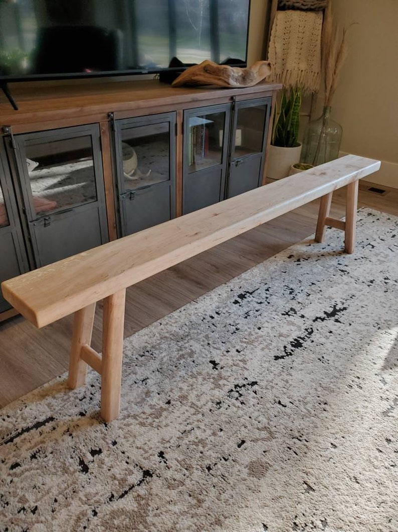May include: A light wood bench with a simple design and two legs on each end. The bench is sitting on a patterned rug in front of a dark wood cabinet with glass doors.