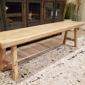 May include: A natural wood bench with a rustic finish and a simple design. The bench has a wide, flat top and two sturdy legs on each side. The bench is sitting on a patterned rug.