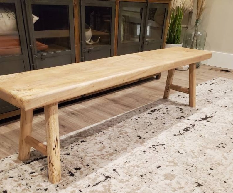 May include: A natural wood bench with a rustic finish and a simple design. The bench has a wide, flat top and two sturdy legs on each side. The bench is sitting on a patterned rug.