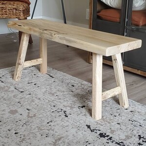 May include: A natural wood bench with a light brown finish and a simple, rustic design. The bench has a wide, flat top and four legs that are slightly angled. The bench is sitting on a patterned rug.