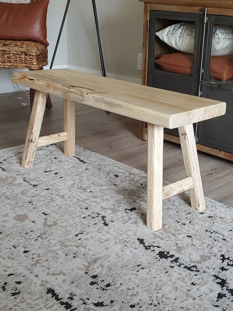 May include: A natural wood bench with a light brown finish and a simple, rustic design. The bench has a wide, flat top and four legs that are slightly angled. The bench is sitting on a patterned rug.