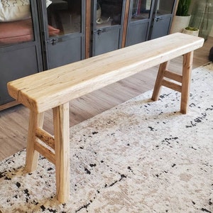 May include: A long, rustic wooden bench with a natural finish and a simple, sturdy design. The bench has two legs on each end, creating a stable and functional piece of furniture.