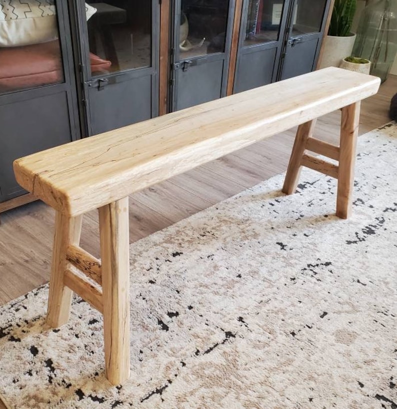 May include: A long, rustic wooden bench with a natural finish and a simple, sturdy design. The bench has two legs on each end, creating a stable and functional piece of furniture.