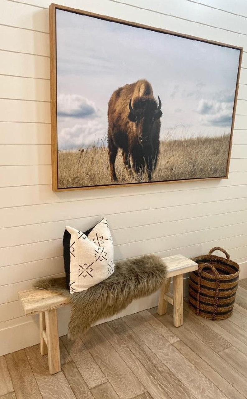 May include: A wooden bench with a faux fur throw and a patterned pillow. A framed print of a brown bison in a field hangs on the wall above the bench. A woven basket sits on the floor next to the bench.