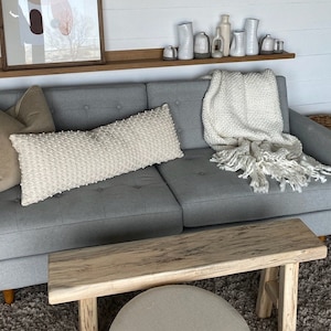 May include: A gray sofa with a white throw blanket and pillows. The sofa is in front of a white wall with a wooden shelf above it. There are white ceramic vases on the shelf. A wooden bench is in front of the sofa.